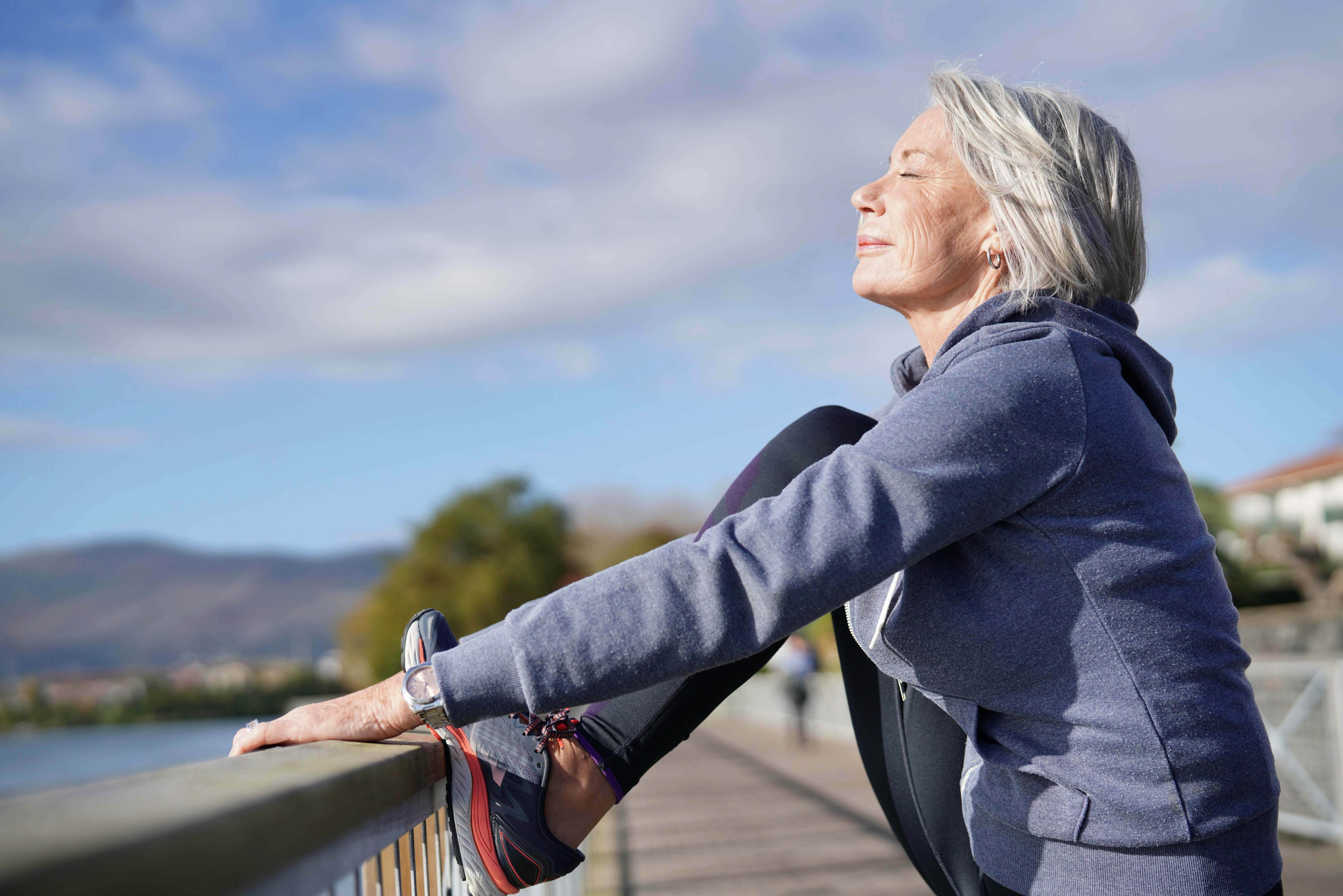 Older woman wearing a hoodie stretching her leg on a barrier near the water, with her face tilted up towards the sun and eyes closed